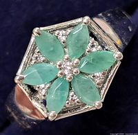 Close-up of silver ring with six marquise-shaped emerald stones arranged in a flower pattern with small accent stones in hexagonal setting.
