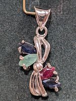 Close-up photo of silver pendant with marquise emerald, ruby, and sapphire stones on a black background.