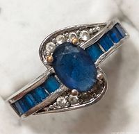 Close-up of the front of the ring showing central oval blue gemstone, small clear stones in heart-shaped design next to the center, and rectangular blue stones set in the band.