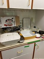 Wide angle photo showing counter with multiple cookware items including food mill, white casserole, yellow casserole dish, and baking trays.
