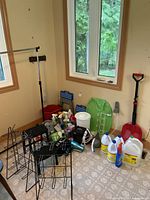 Photo showing laundry racks, cleaning supplies, bucket, iron, hair dryer, step stools, fan, shovel, and other household utility items grouped on floor by two windows.