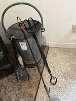 Metal bucket with 3 fireplace tools: shovel, tongs, poker, leaning against wall on carpeted floor
