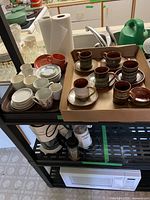 Photo showing box with 8 ceramic mugs and saucers in brown and green glaze, nearby white small plates and 6 floral pattern cups.