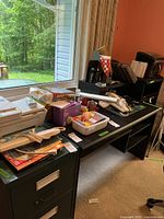 Black desk and filing cabinet near a window with assorted office supplies, snacks, and containers on top.
