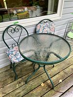 Sets of two green metal chairs with floral cushions and a round glass top table with green metal frame, placed on wooden deck flooring.