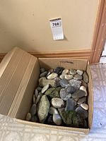 Box filled with numerous assorted rocks and stones of different sizes and colors placed on floor against wall.