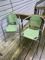 Two light green mesh fabric outdoor chairs with silver metal frames positioned on a wooden deck against a house wall.