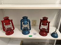 Three vintage metal lanterns on a shelf: two red, one blue, approximately 10 inches tall, plus one small blue oil can with spout and cap.