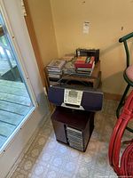 Sony boombox on dark wooden stand next to shelves and stacks of CDs and VHS tapes in storage boxes, placed on floor in corner.