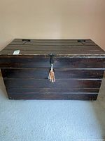 Front view of a rectangular wooden chest with dark wood finish, metal handles on the sides, and latch closure with tassel at center. Surface wear visible.