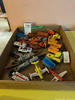 Top-view of cardboard box filled with assorted Matchbox toy vehicles including racing cars, airplanes, battleships, and construction trucks.