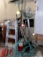 Utility lot items including aluminum step ladder, orange extension cords, green garden hose, fire extinguisher, and multiple yard tools leaning against a wall