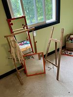 Wooden easel with a weaving loom bar and textile piece, two mirrors leaning against the wall.
