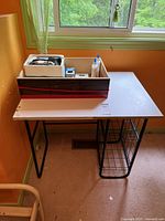 Work desk with wooden top and metal frame with wire basket on side beneath, orange wall background