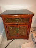 Front view of antique wooden side table with one drawer and double door cabinet below showing decorative gold floral panels and brass hardware.