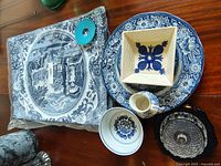 Wide view showing all items in the lot arranged on a wooden surface including porcelain plates, bowls, small pitcher, cup with maker's mark, glass bowl, blue patterned table runner, and small blue ceramic item.