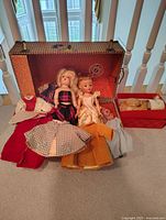 Two vintage dolls inside an open trunk along with several doll clothes including a checkered dress, orange skirt, and light blue skirt. A red garment is also visible.