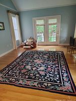 Full room view showing the large rectangular antique needlepoint rug with floral designs placed on hardwood floor beneath natural light from window.