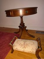 Round antique wooden table with a small drawer and pedestal tripod base, paired with a floral upholstered stool with wooden frame and turned armrests.