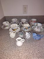 Seven floral decorated teacups and saucers arranged in the back with four uniquely shaped ceramic creamers in front on a kitchen countertop.