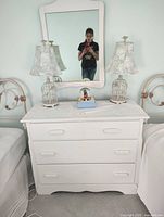 Full view of white vintage solid wood dresser with attached mirror, two birdcage-style lamps, a snow globe and a pastel blue box on top between the lamps