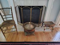 Full lot showing fireplace screen, brass kettle/container with lid, antique wooden chair, and small wooden side table on hardwood floor with rug.