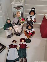 Seven pilgrim and caroler figurines on a white surface near a window, showing standing and seated dolls with detailed clothing and painted faces.