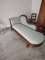 Photo showing the wood-framed chaise lounge with light blue fabric upholstery, curved raised armrest on one side, and carved wooden legs on casters.