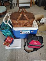 Photo shows a blue wheeled Igloo cooler with white hinged lid, atop the cooler is a woven brown picnic basket with two straps, a red and black Coleman cooler bag, a blue and green patterned soft cooler bag, several Ziploc containers including a white-lid box, and a cardboard box labeled 'Large Bowl'. The items are placed on a concrete floor in a garage space.