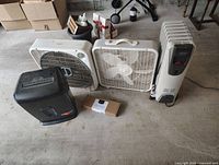 View of two white box fans, one black fan heater, one white oil-filled radiator heater, and one boxed power strip on the floor in a garage.