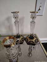 Photo of two ornate glass candlesticks with hexagonal black bases and gold decorations, next to a small faceted glass pitcher with gold handle and rim, displayed on a reflective surface.