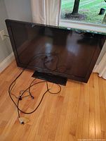 Flat screen TV placed on hardwood floor near window, showing front and base stand with attached cables.