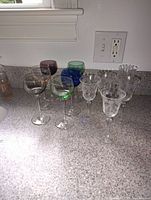 Photo of 10 assorted stemmed glasses on a countertop, including clear and colored glasses in blue, green and purple.
