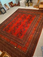Full view of the large red rug showing intricate geometric patterns and multiple borders on a light carpeted floor surrounded by household items.
