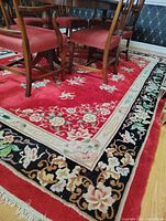 Image showing part of the dining room rug under dining chairs, highlighting red central field with floral motifs and dark floral border with fringe edges.