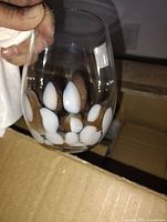 Close-up photo of a single stemless wine glass held by a hand showing the white and gold drop-like design on clear glass.