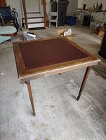 Vintage wooden card table with dark brown felt top and foldable legs, photographed in a garage setting.
