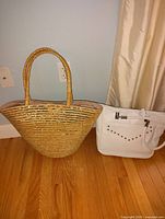 Photo showing two handbags on wood floor against beige wall and curtain: a large woven straw tote and a white Alessia handbag