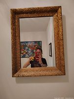 Front-facing photo of the vintage bathroom mirror hanging on a white wall, showing detailed ornate gold frame and reflective glass.