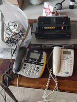 Vintage landline phones, black Linksys device, black modem/router, speaker component, and a bundle of cords on a wooden table.