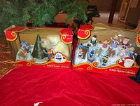 Two vintage Rudolph holiday figurine sets in original red cardboard boxes with plastic windows, showing multiple colorful characters including Rudolph, Bumble, Hermey, and others arranged in festive display.