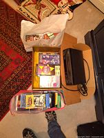 Overhead shot of a black VHS VCR player, a cardboard box filled with VHS tapes, a white paper bag with additional tapes, and a tote bag with more VHS tapes visible inside, on carpeted floor with a patterned rug nearby.