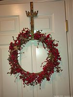 Photo showing a circular wreath made of red berries, brown twigs, and stars, hanging on a metal crown-shaped vertical door hanger against a white door.
