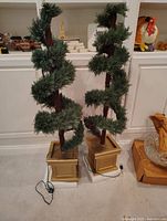 Two artificial Christmas trees with spiral green foliage on brown stems in gold square planters, with cords visible, shown indoors on carpeted floor.