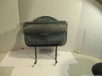 Front angle view of the dark metal mailbox showing embossed leaf and vine decorations and two metal support legs.