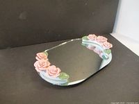 Top angled view showing the mirror’s ceramic rose decorations on two opposite corners with green leaves, clear reflective surface, and smooth edges.