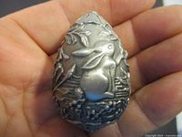 Close-up of pewter egg held in hand showing rabbit relief design and plants.