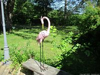 Pink metal flamingo garden decoration standing on wooden railing outdoors, surrounded by greenery and visible lamp post.