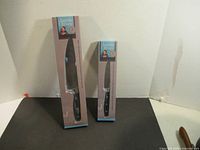 Two boxed Jamie Oliver knives: a larger carving knife and a smaller utility knife, standing upright in their original packaging.