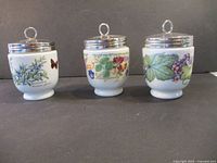 Three porcelain egg coddlers with silver lids displayed side by side showing botanical designs.